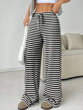 A female modeling the front of a pair of Black Orvella LumaTide Striped Wide Leg Pants paired with a grey short sleeved blouse, jewelry, a white purse, and casual camel loafers.