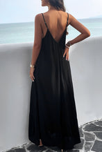 A model showcasing the back of a black Backless Maxi Cami Dress with Pockets paired with jewelry and clear heels posing on an Oceanview balcony 