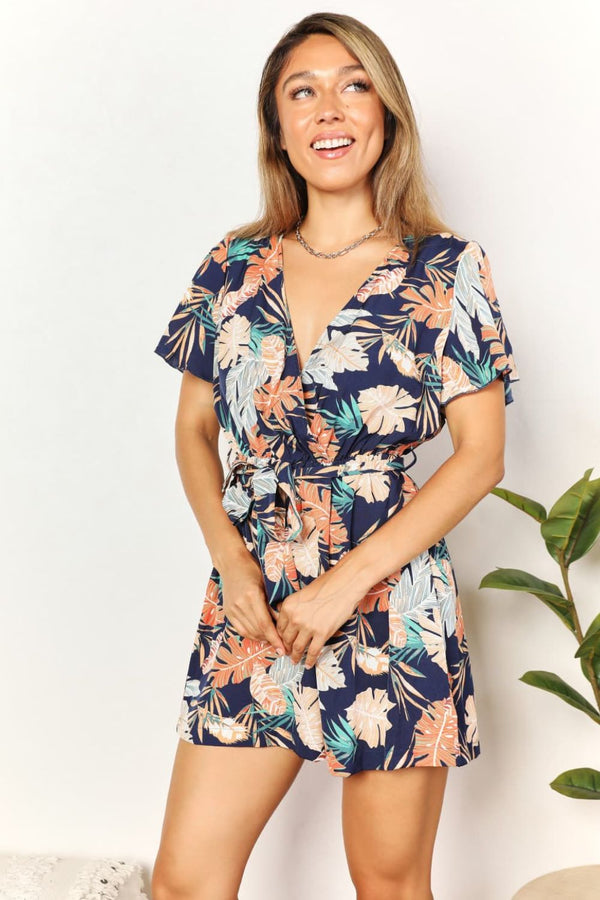 Model wearing botanical print surplice neck romper front view, showing V-neckline, tie waist, and flutter sleeves for a flattering summer look.