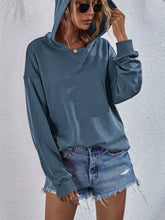 a model wearing a dusty blue Zephyra Breeze Dropped Shoulder Hoodie paired with tinted glasses, jewelry, and light blue denim shorts highlighting drop shoulder seam.