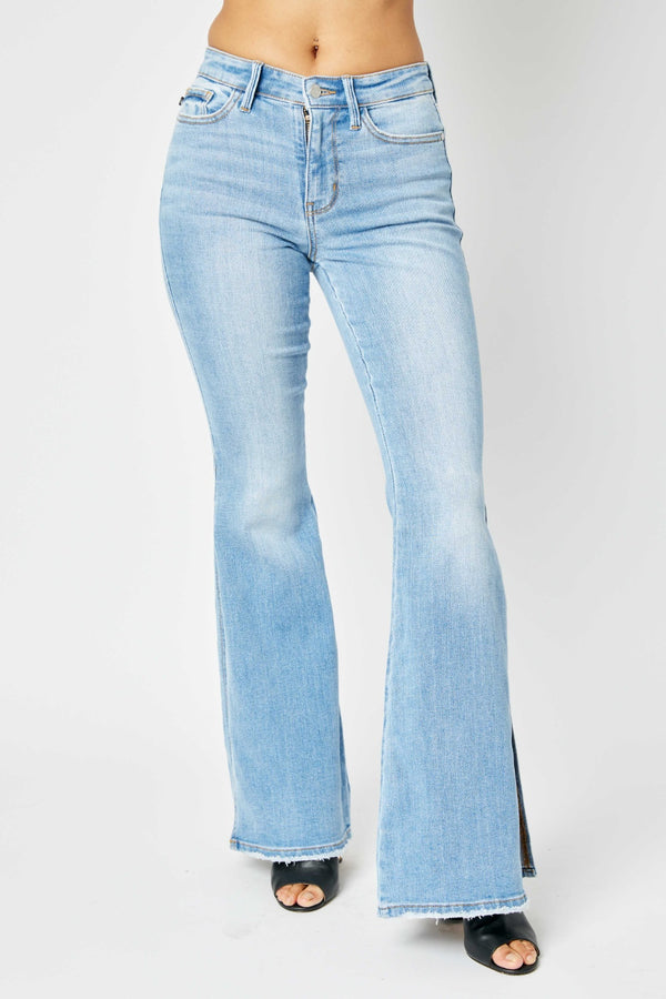 A female posing from the front wearing the light meduim blue Judy Blue Full Size Mid Rise Raw Hem Slit Flare Stonewashed Jeans paired with a red ruffled bloues, jewelry, and black round open-toe heels.