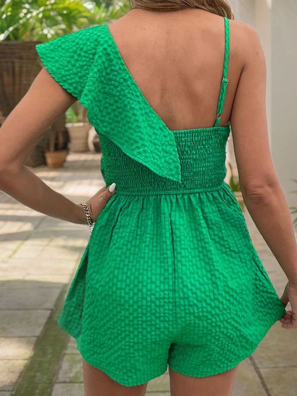 Back view of model wearing green Cymoré Muse Smocked Back Asymmetrical Ruffled Romper, highlighting the smocked back detail, adjustable strap, and relaxed ruffled hem, styled with tan sandals and accessories.