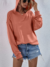 a model wearing a tangerine Zephyra Breeze Dropped Shoulder Hoodie paired with tinted glasses, jewelry, and light blue denim shorts.