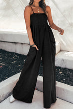 A front photo of the model showcasing  pockets of a black Smocked Amphitrite's Flow Wide Leg Jumpsuit paired with gold jewelry and white casual boots