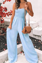 A front photo of the model showcasing  pockets of a misty blue Smocked Amphitrite's Flow Wide Leg Jumpsuit paired with gold jewelry, a brown shoulder purse, and casual boots