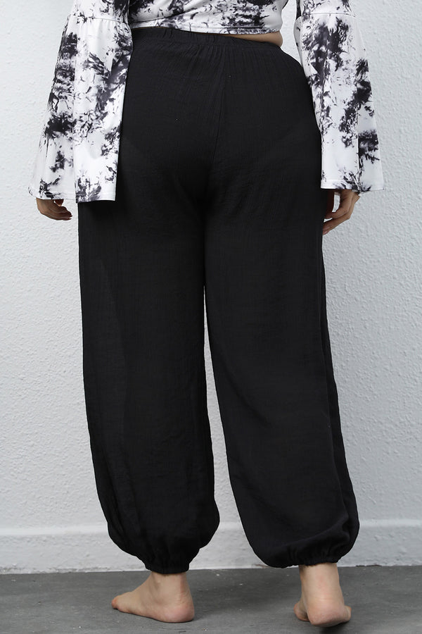 A woman modeling the black of a pair of Auralyn CurveCraft Plus Size Drawstring Jogger Pants paired with a tye-die blouse.