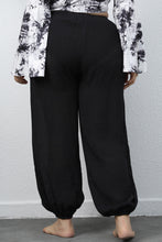 A woman modeling the black of a pair of Auralyn CurveCraft Plus Size Drawstring Jogger Pants paired with a tye-die blouse.
