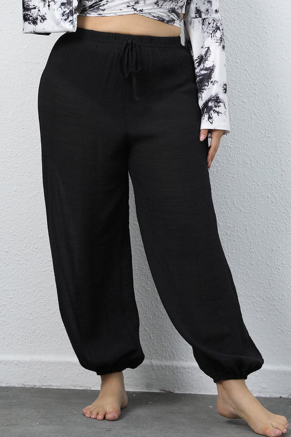 A woman modeling the front side of a pair of black Auralyn CurveCraft Plus Size Drawstring Jogger Pants paired with a tye-die blouse.