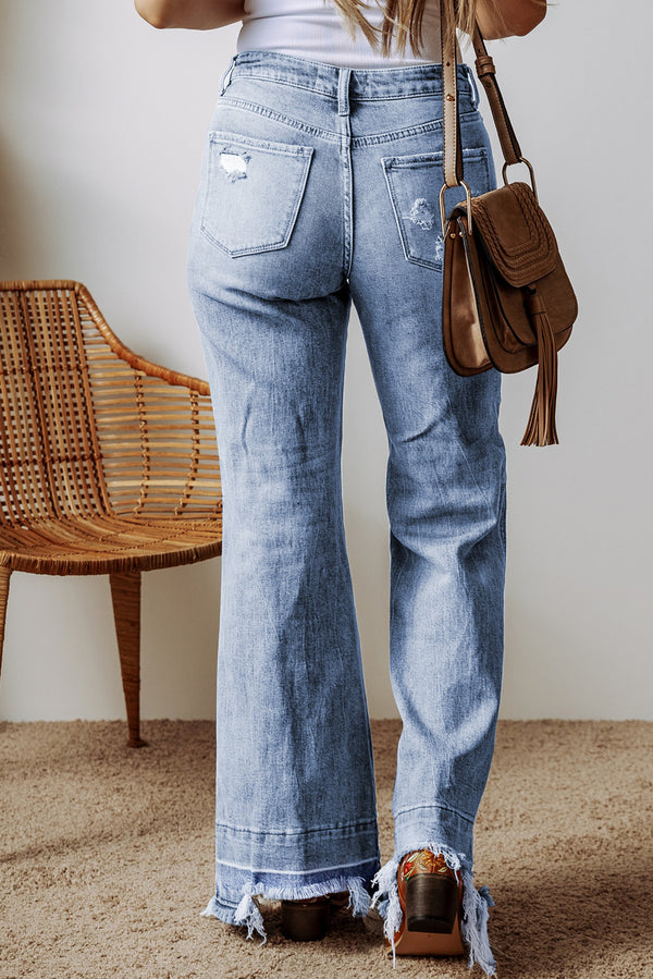 A female posing from the back wearing light medium blue Avaris DriftCrest Raw Hem Bootcut Denim Jeans paired with a white blouse, jewelry, a brown shoulder bag, and brown multicolored loafers.