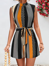 Model wearing the GeoChic Harmony Tied Color Block Sleeveless Romper in black with earth-tone color block design, mock neck, and waist tie—styled with gold hoops and block sandals for a sleek, modern look.