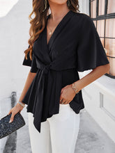 Model standing by a cozy cottage in a  black tied front peplum blouse and denim shorts, perfect for a relaxed weekend getaway vibe.