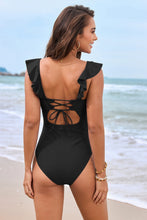 A woman showcasing the back of a black Lisianthus Ruffled Plunge One-Piece Swimsuit standing by ocean waves at Playa del Amor beach.