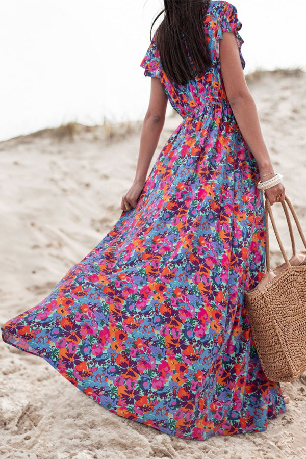 Back view of Bloomirra Slit Floral Print Cap Sleeve Maxi Dress in floral print, model walking along the beach with a woven handbag.