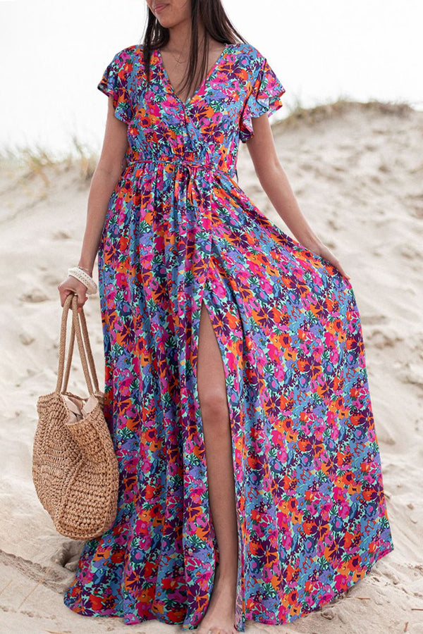 Model wearing Bloomirra Slit Floral Print Cap Sleeve Maxi Dress in floral print, standing on a sunlit beach holding a woven handbag.