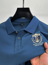 Close-up front view of blue Equestrian Crest Designer Polo on hanger showing embroidery, buttons, and flip color-contrast collar.