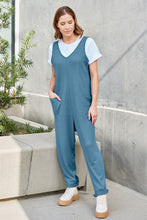 Model overlooking mansion balcony in Sky Blue Caeloura Jumpsuit, beside a tall pine tree at sunset.