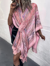 Lifestyle image of model wearing a carnation pink Heathered Fringe Hem Poncho with pom pom fringe, walking through a city park with autumn leaves underfoot.