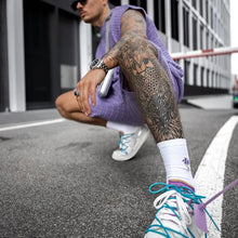 Man in dark violet Draynoir FlexKnit Set in squatted pose with colorful sneakers, accessories.
