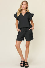 Model lounging in black Facts Only Fit Set with flat sandals, holding iced coffee.
