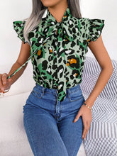 Model styled in a green leopard tie-neck blouse, seated at a rooftop patio table with gold hoop earrings and sleek trousers.