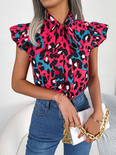 Model wearing a hot pink leopard print tie-neck blouse with cap sleeves, paired with black jeans and a stylish smile, exuding confidence.