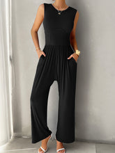 A model showcasing the pockets of a Black color Devine Round Neck Sleeveless Wide Leg Jumpsuit paired with gold jewelry and white heels