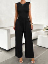 A model posing in the black color Devine Round Neck Sleeveless Wide Leg Jumpsuit paired with gold jewelry, and white heels