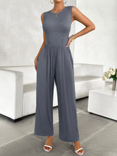 A model posing in the blue-gray color Devine Round Neck Sleeveless Wide Leg Jumpsuit paired with gold jewelry, and white heels