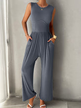 A model showcasing the pockets of a Blue-Gray color Devine Round Neck Sleeveless Wide Leg Jumpsuit paired with gold jewelry and white heels