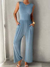 A model showcasing the pockets of a Light Blue color Devine Round Neck Sleeveless Wide Leg Jumpsuit paired with gold jewelry and white heels