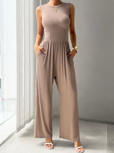A model showcasing the pockets of a Mocha color Devine Round Neck Sleeveless Wide Leg Jumpsuit paired with gold jewelry and white heels