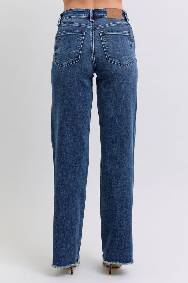 A female posing from the back wearing dark blue Judy Blue Full Size Raw Hem Mid Rise Denim Jeans paired with a red blouse, jewelry, and red pointed-toe heels.