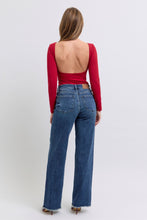 A female posing from the back wearing dark blue Judy Blue Full Size Raw Hem Mid Rise Denim Jeans paired with a plunging back red blouse, jewelry, and red pointed-toe heels.
