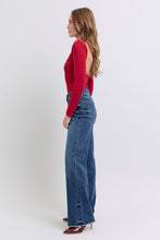 A female posing from the left side wearing dark blue Judy Blue Full Size Raw Hem Mid Rise Denim Jeans paired with a red blouse, jewelry, and red pointed-toe heels.