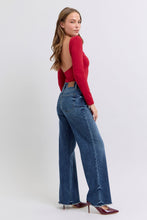 A female posing from the right side wearing dark blue Judy Blue Full Size Raw Hem Mid Rise Denim Jeans paired with a red blouse, jewelry, and red pointed-toe heels.