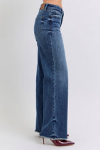 A female posing from the side wearing dark blue Judy Blue Full Size Raw Hem Mid Rise Denim Jeans paired with a red blouse, jewelry, and red pointed-toe heels.