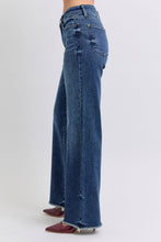 A female posing from the side wearing dark blue Judy Blue Full Size Raw Hem Mid Rise Denim Jeans paired with a red blouse, jewelry, and red pointed-toe heels.