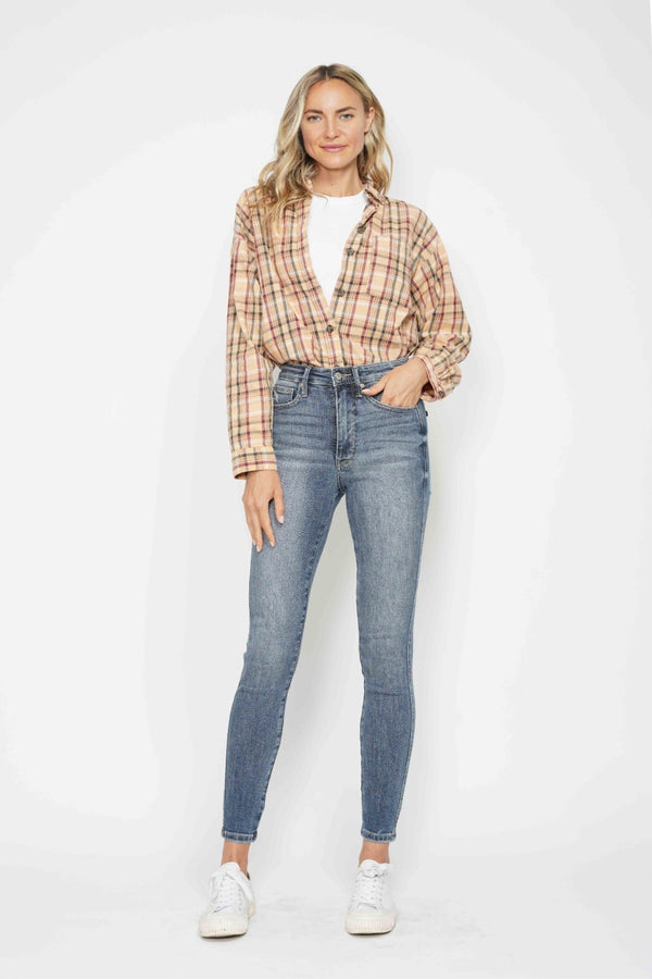 A female posing from the front wearing medium blue Judy Blue Full Size Tummy Control Contrast washed Skinny Jeans paired with a plaid button-up shirt, jewelry, and white sneakers.