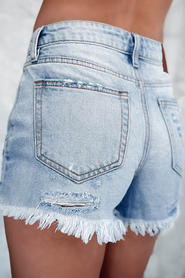 Back view of woman in light blue distressed high waist denim shorts, showing frayed raw hem and pocket details.