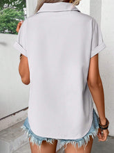 Back view of model showcasing the lightweight, relaxed fit of the light gray pleated collared neck short sleeve blouse, paired with white trousers.