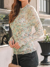 Model wearing light green mock neck thumb holster top, lounging in a cozy coffee shop setting with layered necklaces and crossbody bag.