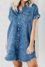 Side-front photo of woman posing  wearing medium blue Oressina Pocketed Raw Hem Denim Dress styled with gold jewelry with white brick wall background.