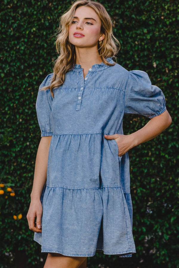 Model front view wearing ODDI Washed Tiered Mini Denim Dress styled with hoop earrings and strappy sandals for a casual daytime look with topiary bush background. 