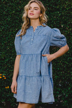 Model front view wearing ODDI Washed Tiered Mini Denim Dress styled with hoop earrings and strappy sandals for a casual daytime look with topiary bush background. 