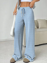 Female model in Light Blue Orvella LumaTide Striped Wide Leg Pants with a white short-sleeved blouse, jewelry, white purse, and camel boots, front view.