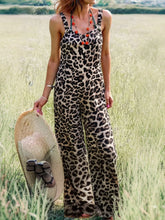 Model standing in a grassy meadow wearing Panthorlisa Leopard Overalls styled with a wide straw hat tied with a bow, layered gold and red beaded jewelry, and stacked gold bangles.