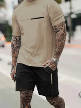 Person wearing a sand Parivor FutureFlex Set; t-shirt and black shorts with tattoos on arms and legs, standing outdoors.