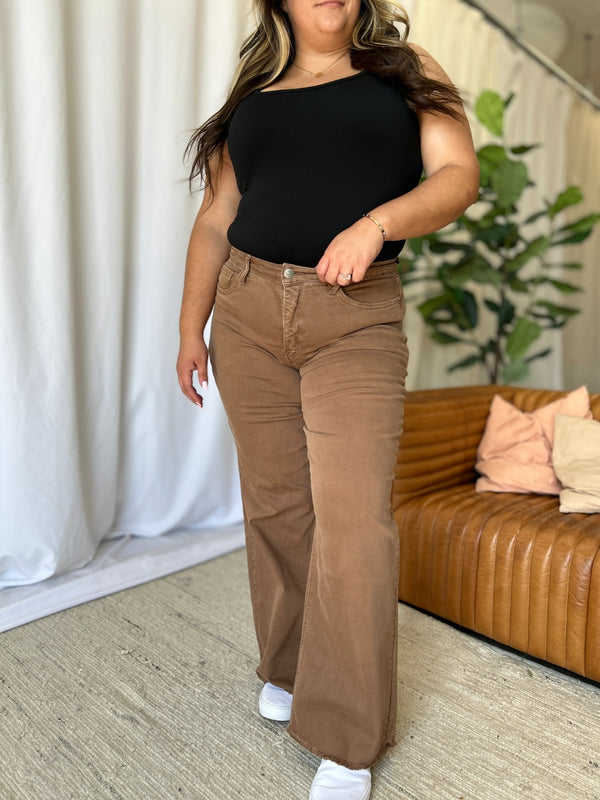 A female modeling the front of coffee brown Full Size High Rise Garment Dye Wide Leg Jeans paired with a black short sleeved blouse, jewelry, and white sneakers.