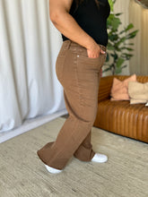 A female modeling the side of a pair of coffee brown Full Size High Rise Garment Dye Wide Leg Jeans paired with a black short sleeved blouse, jewelry, and white sneakers.