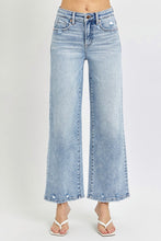A female posing from the front wearing light blue RISEN Full Size Tummy Control High Rise Crop Wide Leg Jeans paired with a floral white blouse, jewelry, and white round open-toe heels.
